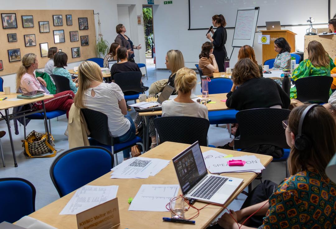 Academy 2023 - A group of around 15 people sitting around tables in a meeting room, looking to the front and listening to two people presenting in front of a screen and flip chart 
