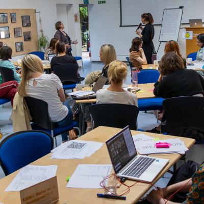 Academy 2023 - A group of around 15 people sitting around tables in a meeting room, looking to the front and listening to two people presenting in front of a screen and flip chart 