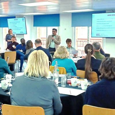 Ayad presenting to a group of about 25 people sitting in chairs in a meeting room. There are 2 large screens in the background with a presentation on