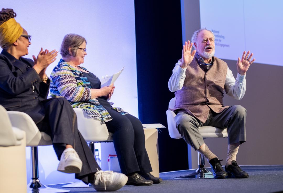 Budd Hall sitting, talking animatedly on stage at Engage with Sophie Duncan and Ima Jackson 
