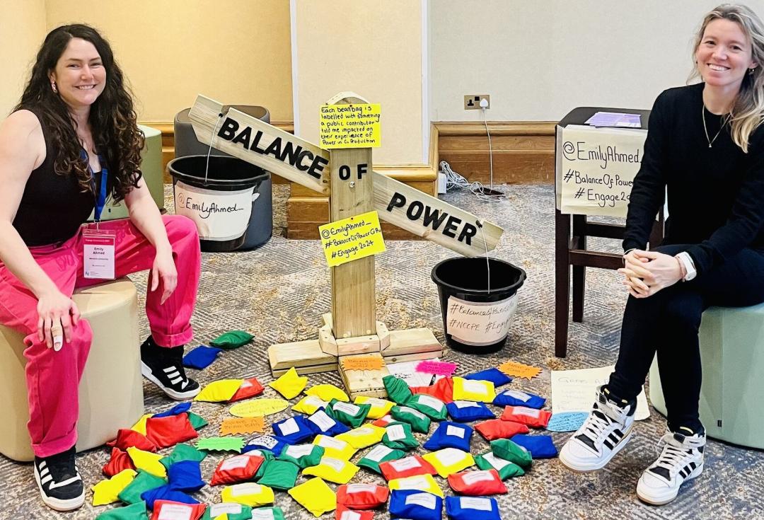 Emily and her colleague sitting on stools either side of Emily's 'balance of power' wooden scales, surrounded by colourful beanbags with labels attached.